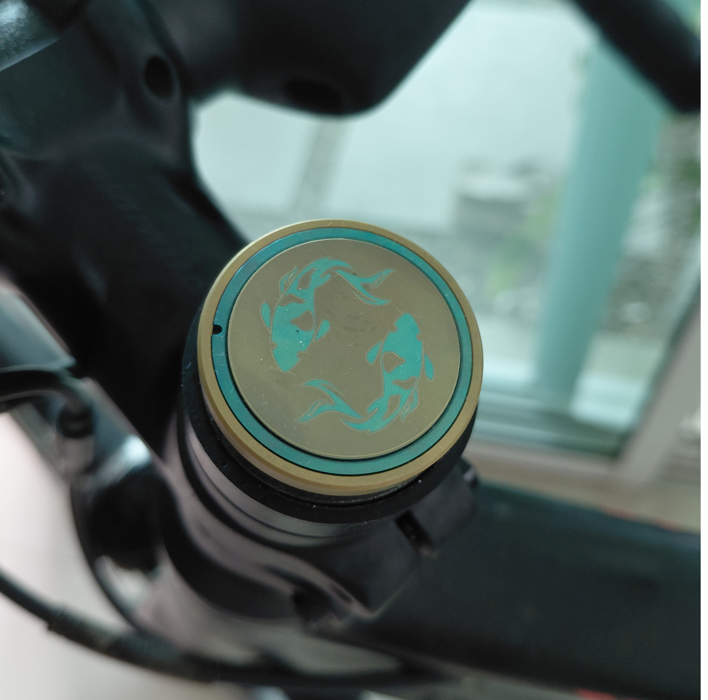 Close-up of a titanium bicycle stem top cap with two Koi designs in turquoise and gold.