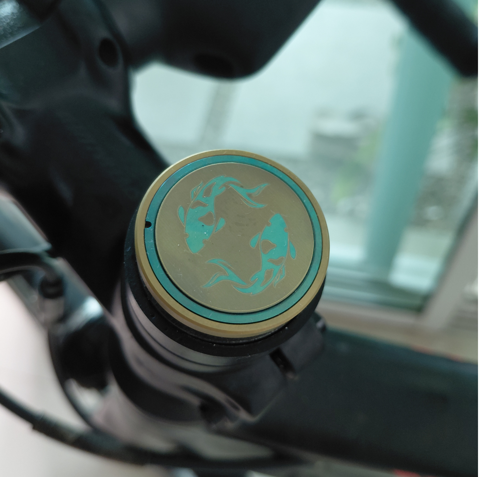 Close-up of a titanium bicycle stem top cap with two Koi designs in turquoise and gold.