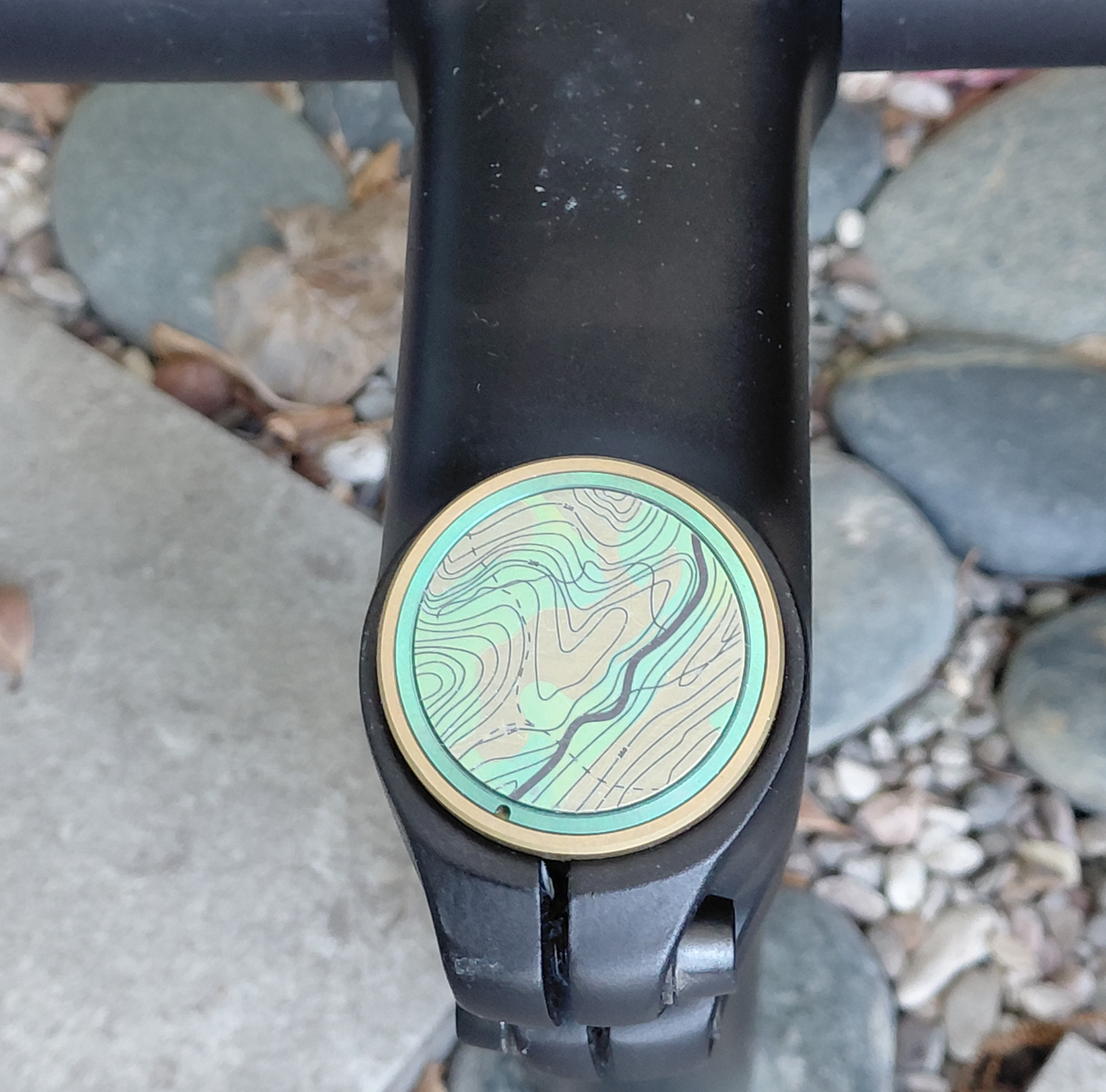Close-up of a titanium bicycle stem top cap with a topographic design on a stone surface.