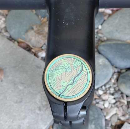Close-up of a titanium bicycle stem top cap with a topographic design on a stone surface.