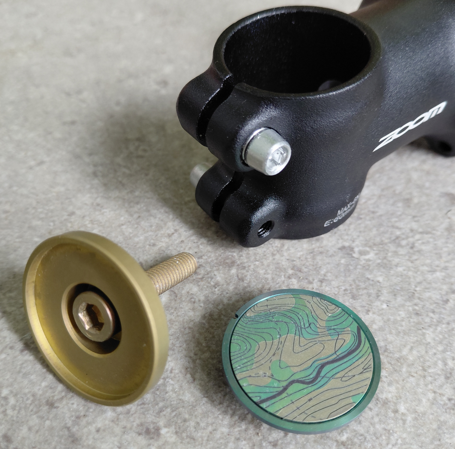 Close-up of a black bicycle stem with gold and green titanium stem top cap on a gray surface.