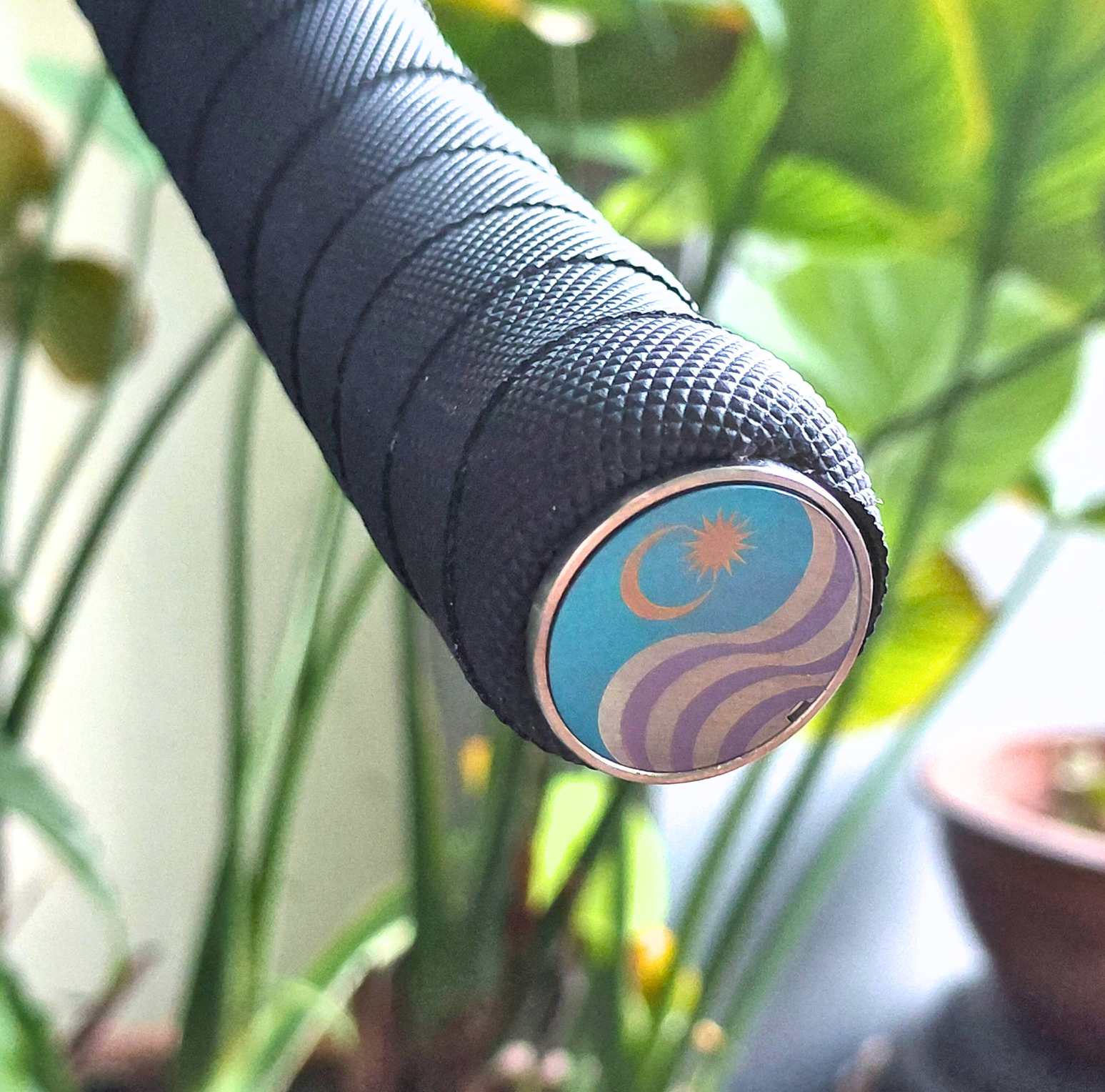 Close-up of a textured black bicycle handle with a blue, gold and purple bicycle End cap  against a blurred green plant background.